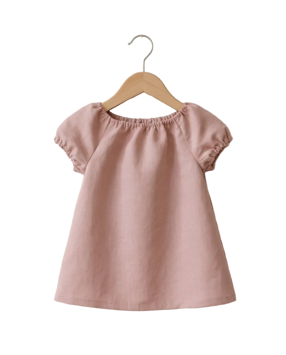 A dusty pink short-sleeve top with a gathered, elasticized neckline, hanging on a wooden hanger against a textured off-white wall.