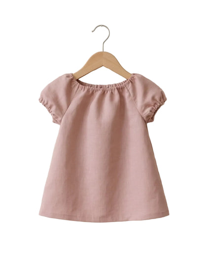A dusty pink short-sleeve top with a gathered, elasticized neckline, hanging on a wooden hanger against a textured off-white wall.